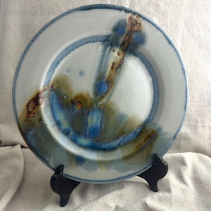 May include: Handmade ceramic plate with a white base and blue and brown abstract design. The plate has a wide rim with a blue border and is displayed on a black stand. The plate's design features a blend of colors.
