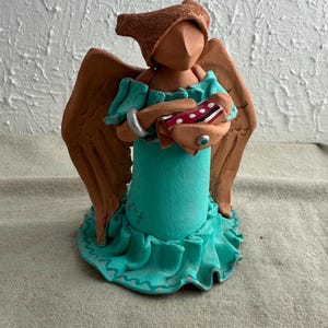 May include: A terracotta angel figurine with wings, wearing a turquoise dress and holding a red and white box. The angel has a brown hat and a silver bracelet. The dress has a ruffled hem and the words "Sharing is Love" written on the front.