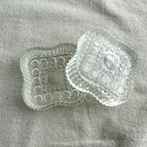 May include: Two clear, vintage glass dishes with lids. The dishes have a rectangular shape with a scalloped edge and a pattern of circles and geometric shapes. The lid is slightly ajar, revealing the interior.