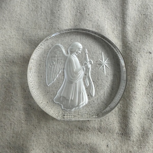 May include: Clear glass angel ornament with a detailed relief of an angel holding a candle and a star. The angel has detailed wings and flowing robes. The ornament is round and has a smooth, polished finish.