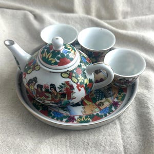 May include: A miniature porcelain tea set featuring a teapot, three cups, and a serving tray. The set is decorated with colorful floral and figural designs, including red, green, and yellow accents. The teapot has a white body and a matching lid.