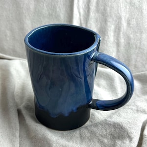 May include: A handmade ceramic mug with a dark blue glaze that fades to black at the base. The mug has a curved handle and a textured surface, perfect for enjoying coffee or tea.