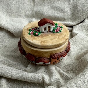 May include: A ceramic trinket box with a lid featuring a miniature house with a red roof and pink flowers. The base of the box is decorated with small houses with red tile roofs. The box is tan and brown.