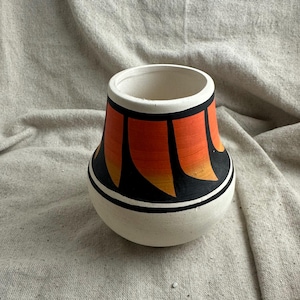 May include: A handmade, off-white ceramic vase with a rounded base and a wide opening. The vase features a striking design of orange and black vertical shapes, accented with black and white bands. A unique decorative piece.