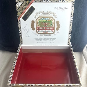 May include: A white wooden box with gold trim and a red interior. The box has a label that reads "Arturo Fuente Imported" and "Double Chateau Fuente Select Long Filler".