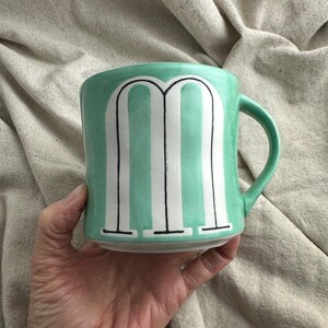 May include: A ceramic mug with a mint green glaze and a white and black striped design. The mug features a large letter "M" in the center. The mug has a curved handle and is held by a hand.