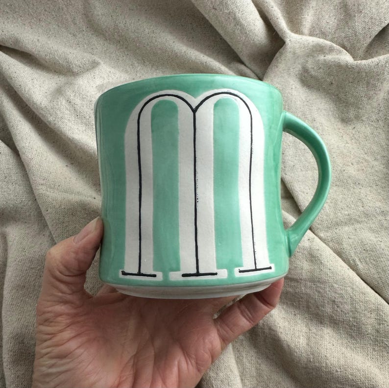 May include: A ceramic mug with a mint green glaze and a white and black striped design. The mug features a large letter "M" in the center. The mug has a curved handle and is held by a hand.