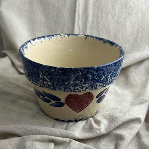 Peut inclure: Un pot de fleur en céramique crème avec un bord bleu moucheté et un motif de cœur rouge. Des accents bleus en forme de feuilles entourent le cœur. Le pot a un aspect rustique et artisanal.