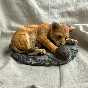 May include: A ceramic figurine of a lion cub with golden-brown fur and dark spots, lying down on a gray rock base. The cub is looking down at a small, round, dark brown textured ball held between its front paws, featuring detailed paw pads.