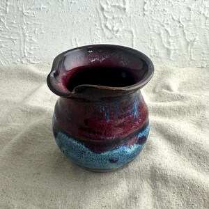 May include: A handmade ceramic pitcher with a dark burgundy interior and rim. The body transitions from burgundy to a vibrant blue, creating a striking contrast. The pitcher has a spout for easy pouring and is a unique piece of functional art.