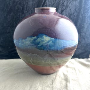 May include: A round ceramic vase with a brown, blue, and green glaze. The vase has a wide mouth and a narrow base.