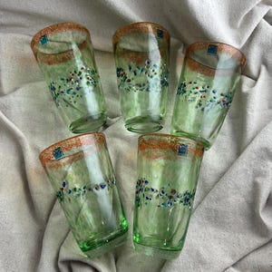 May include: Five green-tinted drinking glasses with a decorative floral pattern in blue, red, and white. The rims are a speckled orange-brown. Each glass has a small square label near the rim. The glasses are arranged on a neutral-colored fabric.