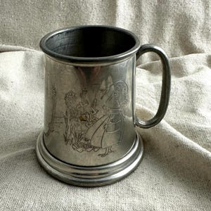 May include: A vintage pewter tankard with a handle and a wide base. The mug features an etched design of figures in a garden setting. The metal has a slightly tarnished appearance, giving it a rustic charm. The mug is designed for holding beverages.