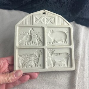 May include: A white ceramic cookie cutter shaped like a barn with four different animal designs: a goose, a pig, a sheep, and a cow. The barn has a red roof and a white cross on the front.