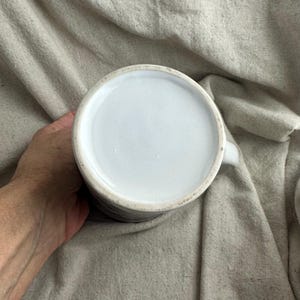 May include: A white ceramic mug with a handle, viewed from the bottom. The interior is white, and the exterior has a dark, textured pattern. The mug is held against a neutral-colored fabric background.