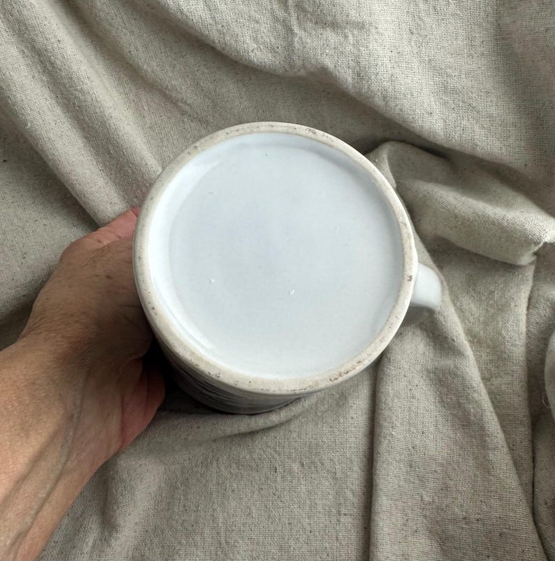 May include: A white ceramic mug with a handle, viewed from the bottom. The interior is white, and the exterior has a dark, textured pattern. The mug is held against a neutral-colored fabric background.