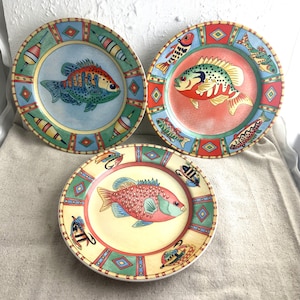 May include: Three ceramic plates with a colorful fish design. Each plate features a different colored fish with a patterned border of fishing lures and bobbers.
