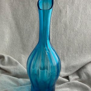 May include: A tall, turquoise glass vase with a flared opening. The vase has a rounded base and a slender neck. Vertical ridges run the length of the vase, creating a textured surface. The vase is approximately 10 inches tall.