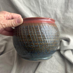 May include: A handcrafted ceramic bowl with a textured, grid-like pattern in shades of brown and blue. The rim is a deep red. The bowl is round and has a small base.