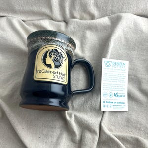 May include: A navy blue Deneen Pottery mug with a handle, featuring a gold label with the text "reClaimed Hair STUDIO". The mug has a dark green and tan rim. A white card with text is next to the mug.