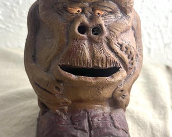 May include: A brown ceramic piggy bank shaped like a cartoon monster with a large nose and a wide mouth. The piggy bank has a slot for coins in the mouth.