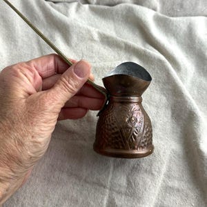 May include: A small, copper-colored Turkish coffee pot with a long, thin handle. The pot has a textured surface with floral designs and a silver-colored interior. The handle is made of a thin, gold-colored metal.