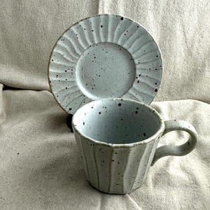 May include: A light blue ceramic teacup and saucer set. The cup has a ribbed design and a handle. The saucer is round with a matching ribbed pattern. Both pieces have dark speckles.