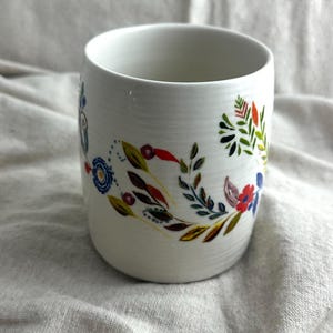 Op de afbeelding: Witte keramische mok met een bloemenpatroon in rood, blauw, groen en geel. De bloemen en bladeren zijn in een decoratief patroon rond de mok gerangschikt. De mok heeft een cilindrische vorm.