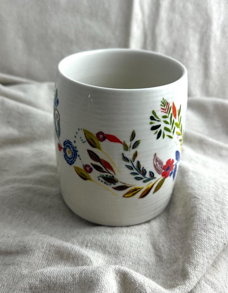 Op de afbeelding: Witte keramische mok met een bloemenpatroon in rood, blauw, groen en geel. De bloemen en bladeren zijn in een decoratief patroon rond de mok gerangschikt. De mok heeft een cilindrische vorm.
