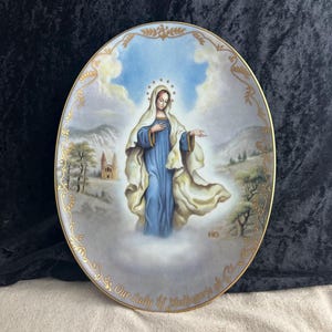 May include: Oval porcelain plate with a gold trim and a depiction of Our Lady of Medjugorje. The image shows a woman in a blue robe with a white veil, standing on a cloud with a church in the background.