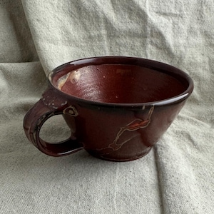 May include: A handmade, maroon-colored ceramic mug with a rounded handle. The mug has a wide opening and a dark brown rim. The interior is a lighter shade of maroon. The mug has a unique, rustic appearance.