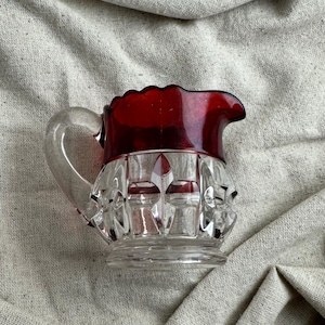 May include: A small, clear glass pitcher with a ruby red rim and handle. The pitcher has a cut glass design on the body and a curved handle. The pitcher is likely used for serving liquids.
