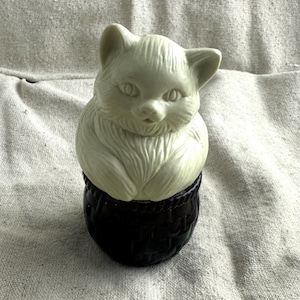 May include: A cream-colored cat figurine atop a dark brown, basket-like base. The cat has detailed fur and facial features. The figurine is set against a neutral, textured background.
