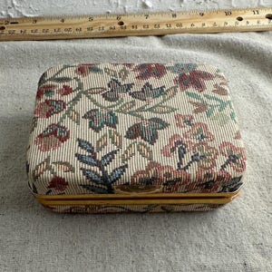 May include: A small, rectangular box with a floral pattern and gold trim. The box is made of fabric and has a hinged lid.
