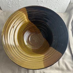 May include: A large, ceramic serving bowl with a black and brown glaze on one half and a yellow and brown glaze on the other half. The bowl has a rippled design.
