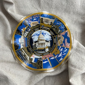 May include: A clear glass decorative plate featuring a central image of the U.S. Capitol Building, surrounded by illustrations of other Washington D.C. landmarks. The plate has a blue and gold border and is set against a neutral fabric background.