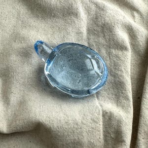 May include: A light blue glass turtle figurine. The turtle has a detailed shell pattern and a small head and tail. The figurine is made of transparent glass and sits on a beige fabric surface.