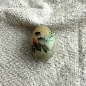 May include: A hand-painted, oval-shaped egg with a light green hue. The egg features a detailed illustration of a bird, red flower, and green foliage. The egg has a handwritten inscription in cursive.