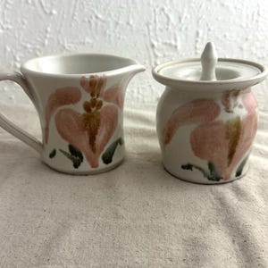 May include: A cream-colored ceramic creamer and sugar bowl set. Each piece is decorated with a pink floral design with brown and green accents. The creamer has a handle and spout, while the sugar bowl has a lid with a small knob.
