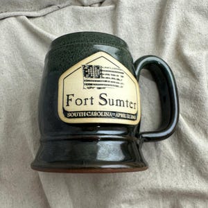 May include: Dark green ceramic beer stein with a black handle. The stein features a cream-coloured label with the text "Fort Sumter" and an American flag illustration. The text "South Carolina, April 12, 1861" is also on the label.
