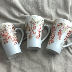 May include: Three white ceramic mugs featuring a hand-painted botanical pattern of red currants and green leaves. Each mug has a tapered cylindrical shape, a smooth glossy finish, and a curved handle extending from the side of the vessel.