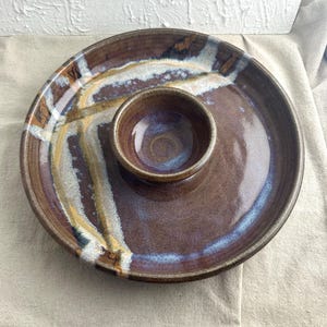 May include: A brown ceramic serving dish with a smaller bowl in the center. The dish has a white and brown glaze with a blue and purple iridescent finish.