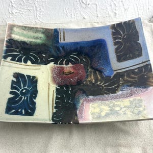 May include: A rectangular ceramic serving dish with a unique, abstract design. The dish features a mix of colors, including blue, black, red, and cream, with floral and geometric patterns. The edges are slightly curved, and the overall aesthetic is artistic.