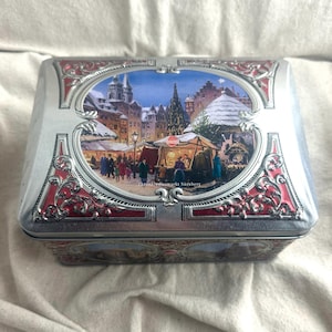 May include: A rectangular silver tin with a detailed illustration of a Christmas market scene. The tin features a red and silver ornate border. The image depicts a festive market with people, stalls, and a Christmas tree.