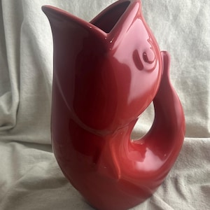 May include: A glossy red ceramic pitcher shaped like a fish with an open mouth at the top, a curved body, and a tail fin forming the handle. The surface is smooth and reflective, featuring molded details for the eye and scales on the side.