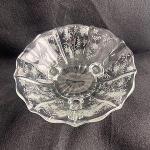 May include: Clear glass serving bowl with a scalloped edge and floral etched design. The bowl has three small feet and a detailed pattern of flowers and leaves. The bowl is empty and sits on a dark surface.