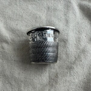 May include: A silver thimble with the text "JUST A THIMBLE" etched around the top. The thimble has a textured, dimpled pattern on the lower half and a smooth, rounded rim.