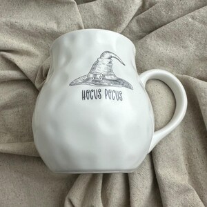 May include: White ceramic mug with a rounded body and a large handle. The mug features a black and white illustration of a witch's hat and the words "Hocus Pocus" in a stylized font. The mug is likely for beverages.