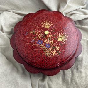 May include: A round, red decorative box with a scalloped edge. The lid features a floral design with gold, blue, and green accents. The box has a textured surface and a small, gold knob.