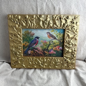 May include: A gold-colored framed picture featuring a painting of two birds perched on branches amidst colorful flowers. The frame has a detailed relief design of figures and cherubs.